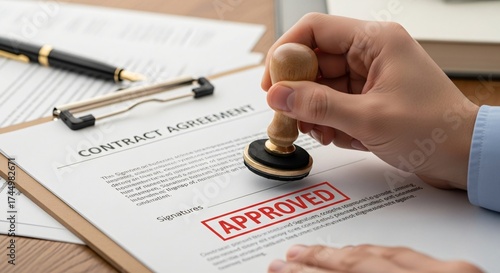 Hand holding a rubber stamp imprinting the word APPROVED onto a contract agreement document