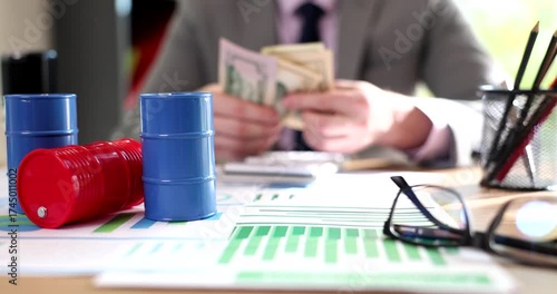 Man counts cash on desk stacking dollar banknotes near blue toy barrels. Company accountant checks amount tracking budget while oil prices rise