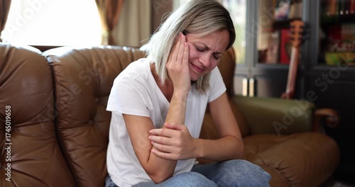 Sad woman suffers from strong toothache sitting on leather sofa in living room. Blonde female holds cheek with hand trying to reduce jaw pain at home
