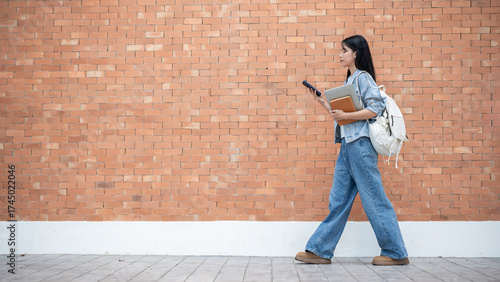 Asian woman student looking at phone holding laptop and book while walking in front of a brick wall.