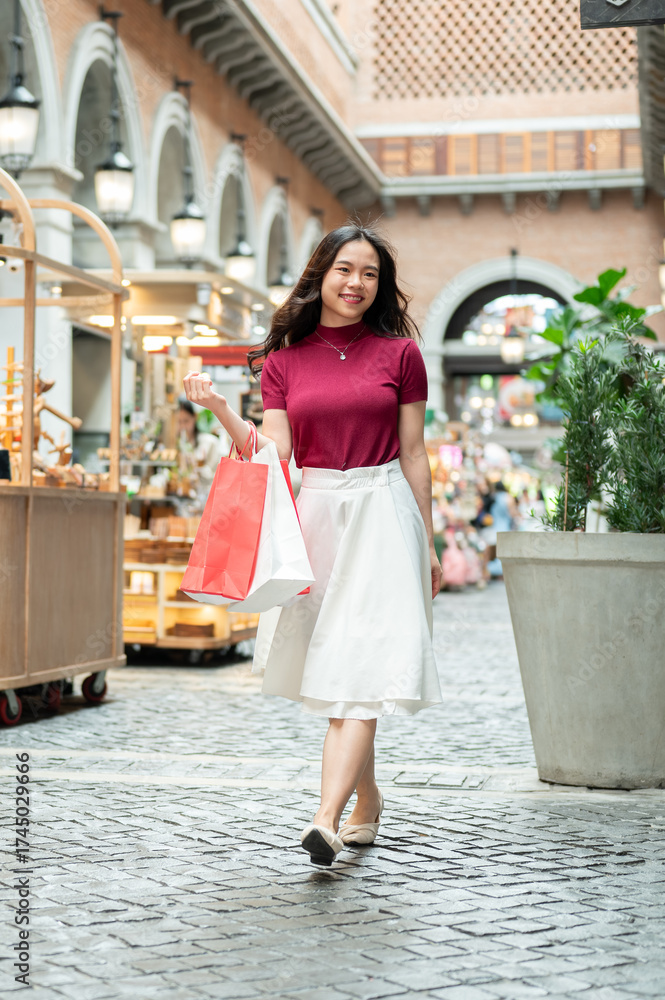 Obraz premium Smiling asian woman holding shopping bags while walking and looking around an indoor market or mall.