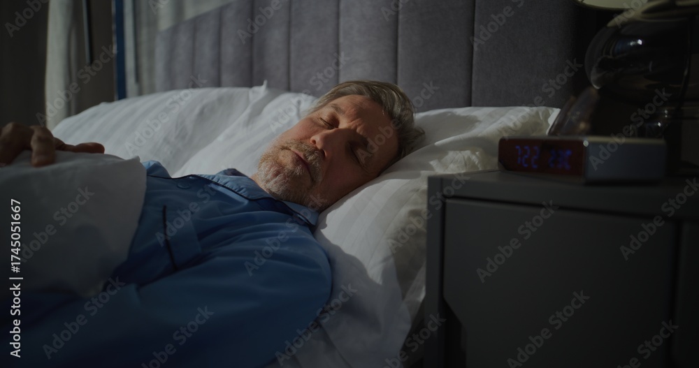 Fototapeta premium Mature Man Slowly Wakes Up Early In Morning In Bedroom Filled With Bright Sunlight. Digital Alarm Clock Shows 7 AM. Slow Morning, Time in Bed. Concept of Morning Routine, Good Morning. Static Shot.