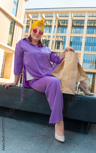 woman with craft shopping bags in the city. The concept of fashionable Consumerism and ecology. An adult Asian woman in a purple suit with red and yellow accents enjoys shopping . a sunny day.