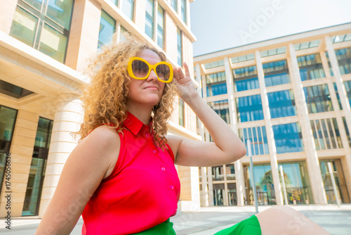 Young blonde woman with long curly hair in red and green fashion closes in sun glasses playful smile happy positive adjusts his glasses