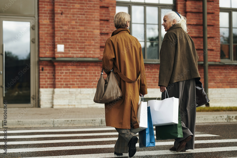 Naklejka premium Two senior Caucasian women walking across crosswalk carrying shopping bags and handbags, engaging in conversation, urban background with brick building visible behind them