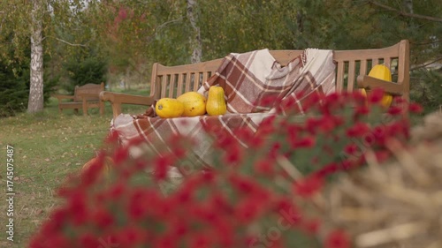 Discover the serene beauty of a cozy wooden bench draped with a soft blanket, decorated with pumpkins, enveloped by a vibrant garden of flowers and towering trees in autumn's embrace.