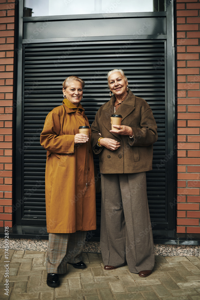 Naklejka premium Two senior Caucasian women standing outdoors holding takeaway coffee cups, smiling and looking at camera, wearing autumn jackets, standing in front of modern brick building