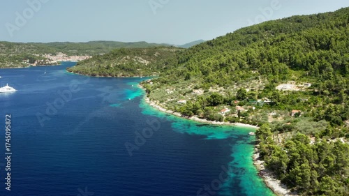 A beautiful Croatian bay with deep blue and turquoise water surrounded by a lush pine forest. A luxury yacht is anchored in the calm sea near a small coastal village in this wide aerial view.