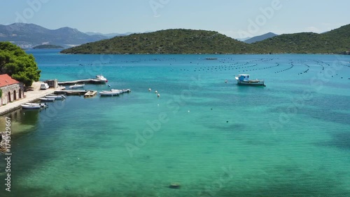 Aquaculture farm for growing oysters and mussels in the clear turquoise water of the Adriatic Sea. A serene view of the Croatian coast with fishing boats near a pier and green hills.