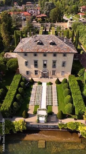 Aerial drone view of Villa Balbiano on Lake Como during a luxury outdoor wedding with floral arch and rows of white chairs on green lawn, overlooking panoramic lake and mountain views