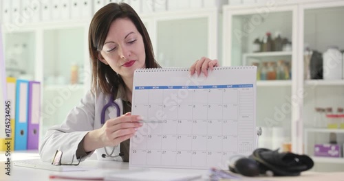 Woman doctor in white lab coat holds up calendar and points at free date. Physician shows available slot for appointment in office. Slow motion