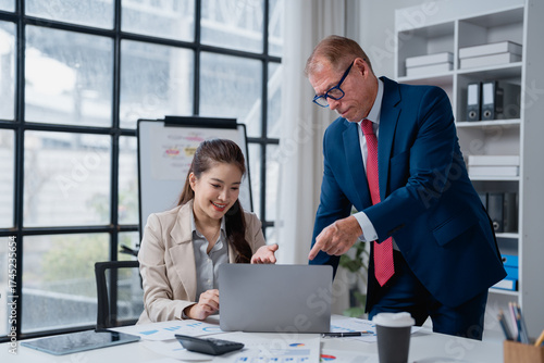 Experienced senior executive mentoring young female colleague. Pointing at laptop screen. Discussing business strategy. And analyzing financial data on documents in a collaborating office environment