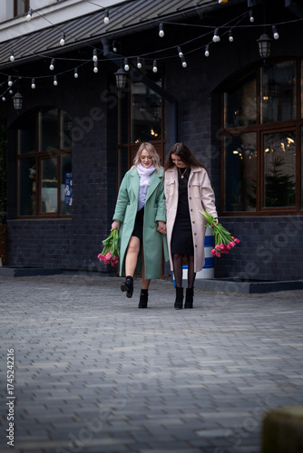 Two women walking together in stylish coats, holding vibrant bouquets of flowers, enjoying a cheerful moment outdoors, showcasing friendship and joy in a lively urban setting