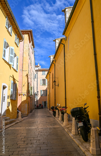 A charming narrow street in Saint-Tropez lined with pastel buildings, shutters, and parked scooters under a bright blue Mediterranean sky