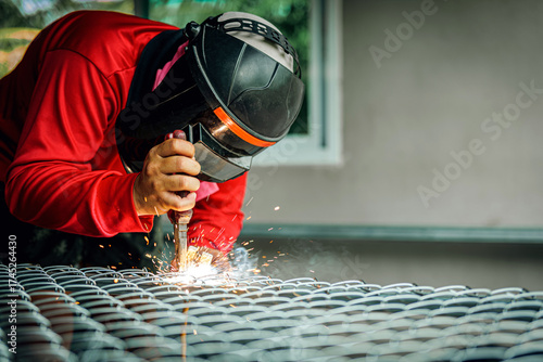 Welder wearing a red long-sleeved shirt and a protective mask is welding a steel grating, Industrial Welder With Torch