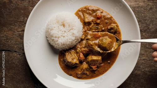 A plate holds white rice and a stew, a spoon rests in the stew, hand present