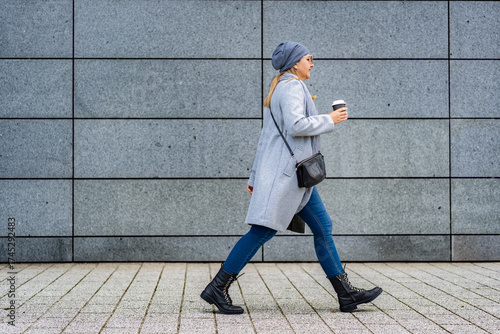 Mature woman walking against background of gray modern wall in autumn. Copy space. Side view