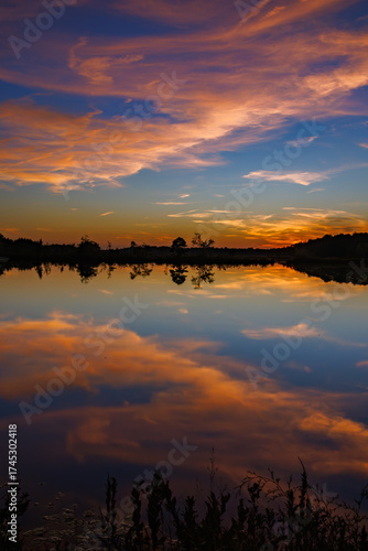 Brilliant sunset sky with vivid orange and pink clouds mirrored in the still waters of a Pine Barrens lake during a calm and peaceful evening.