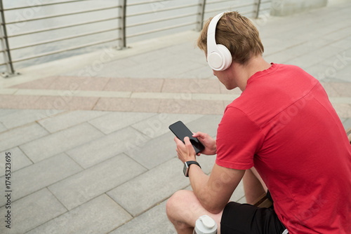 Young man sitting outdoors wearing headphones using smartphone holding device with both hands looking at screen water bottle placed beside on steps