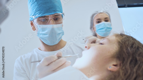 A dentist in blue scrubs and a mask examines a patient's mouth with a dental tool. The patient, wearing a blue mask, sits in a dental chair.