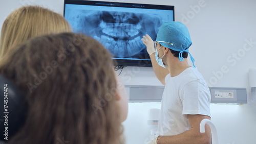 A dentist in a white uniform and blue cap explains dental X-ray results to attentive patients in a modern clinic, emphasizing dental health education in a professional.