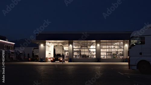 A lone mechanic works late in a well-lit auto repair shop, surrounded by parked trucks. The scene highlights the dedication and precision of a maintenance engineer.