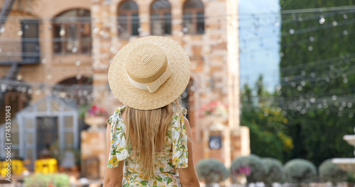 Woman in summer dress and straw hat admires view of rustic courtyard, stone building, fairy lights. Concept: travel, leisure, European architecture.