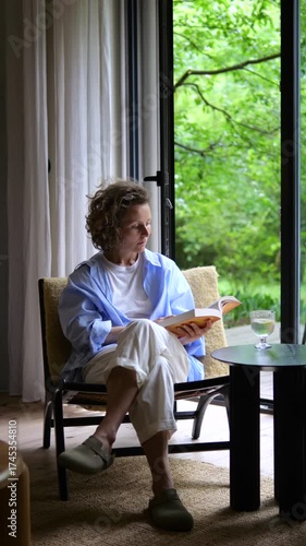 Thoughtful middle aged woman with curly hair sitting in a cozy armchair near a large window, enjoying a relaxing moment while reading a captivating book in her modern living room