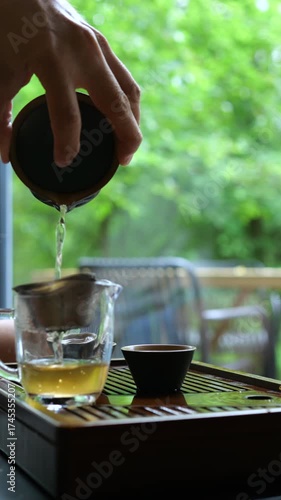 Man's hand pouring hot freshly brewed green tea from a gaiwan through a strainer into a glass pitcher during a traditional chinese tea ceremony, with a lush green garden in the background