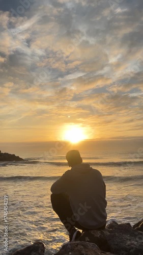 A man sits on a rock overlooking the ocean, watching the sun set. The sky is filled with clouds, creating a moody atmosphere. The man is lost in thought