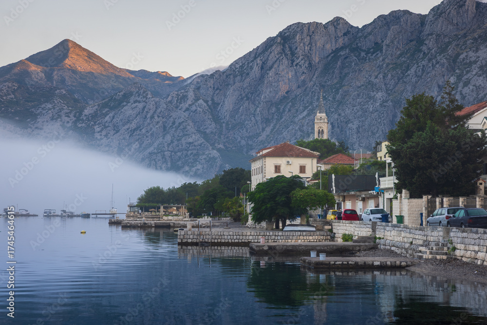 Fototapeta premium Scenic view of Dobrota, Montenegro in fog, with traditional Mediterranean houses, church tower, and mountains rising above the calm Bay of Kotor on a sunny day.