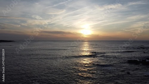 Aerial drone footage flying over the ocean at sunset, capturing golden reflections on the water, gentle waves, and a serene horizon with a colorful twilight sky  
