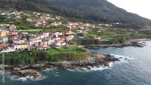 Aerial drone footage of a coastal village with colorful houses, lush green fields, rugged cliffs, and waves crashing on rocky shores, surrounded by forested hills and serene landscapes  
