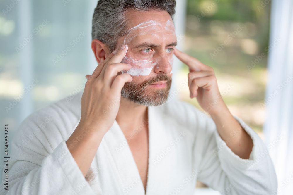 Fototapeta premium Middle-aged man applying facial cream during a morning skincare routine at home. Mature man using a moisturizing mask for anti-aging and rejuvenation. Male cosmetic facial treatment.