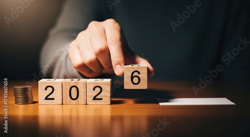 A hand is changing the number on wooden blocks from 2026, with coins nearby, time, finance, planning for the future.