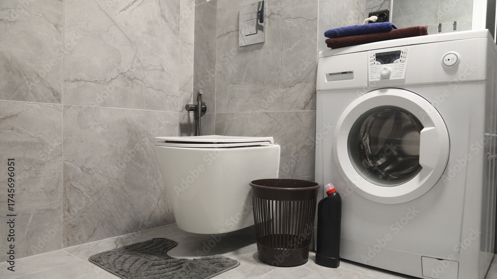 Fototapeta premium Contemporary bathroom interior with wall-mounted toilet, washing machine, laundry basket and detergent on grey tiled background; clean home design and practical lifestyle concept