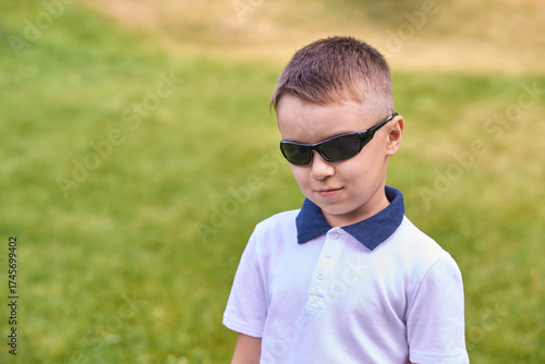 Confident caucasian young boy in sunglasses standing on grass looking cool in white shirt.