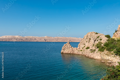 The stunning rocky shoreline of the Croatian coast meets the tranquil, deep blue Adriatic Sea on a bright, sunny day, with the majestic, arid mountains of Pag island on the horizon near Karlobag.