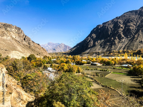 A peaceful village rests beneath the towering Karakoram Mountains, with a flowing river and vibrant autumn trees creating a stunning natural setting