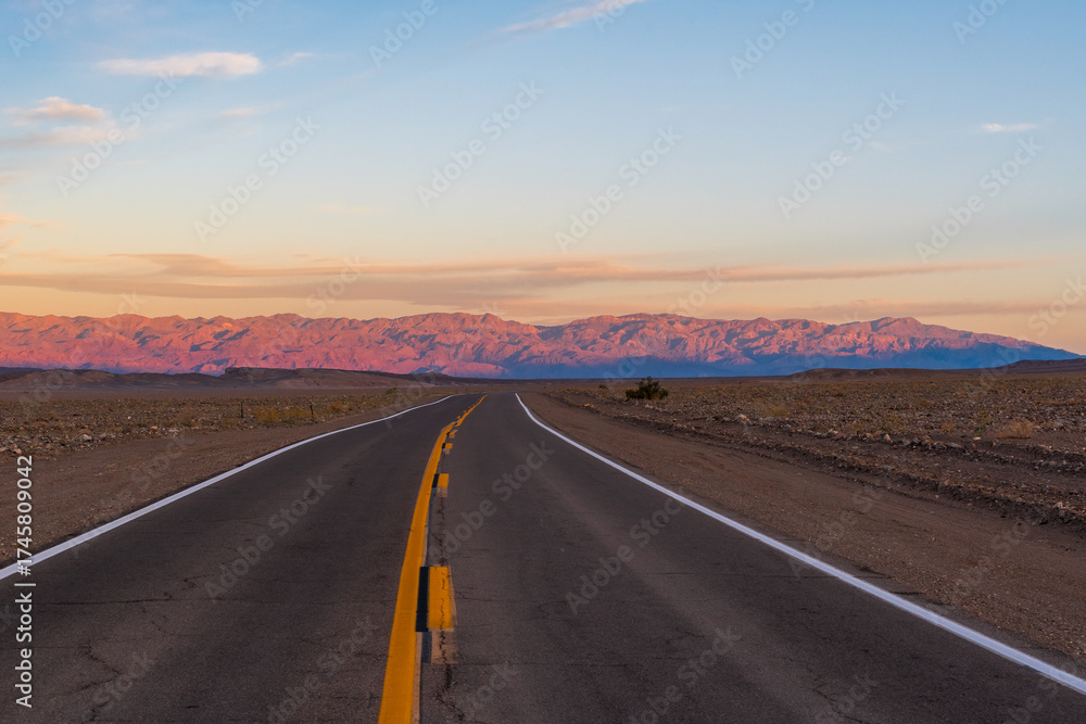 Naklejka premium Desert Road Leading to Pink-Lit Mountains Under Pale Blue Sky with Pink-Tinged Clouds, Death Valley National Park