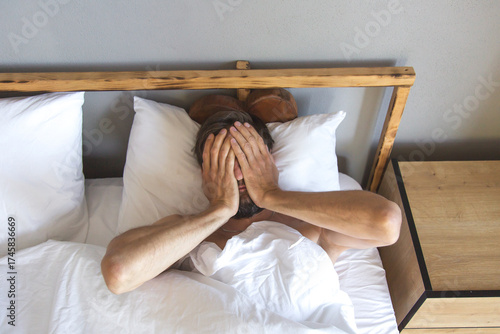 Bearded man lying in bed covering his face with hands after waking up, expressing tiredness and reluctance to get up, concept of morning mood, rest, laziness and everyday routine at home