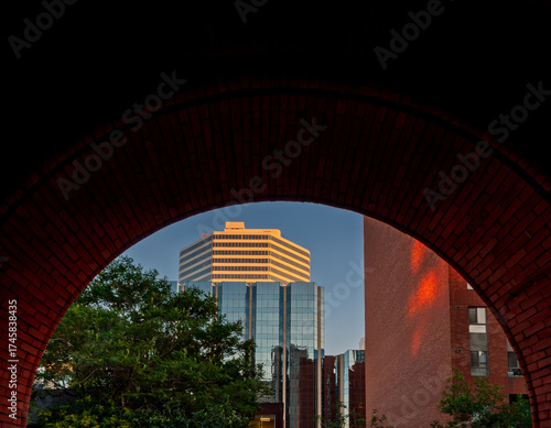  A prominent high-rise modern building with reflective glass panels viewed from and framed by a tunnel-like red brick archway at sunset