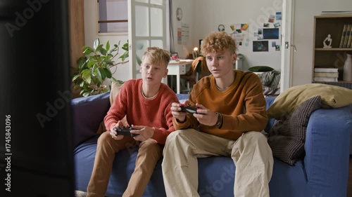 Front view shot of two teenage boys as brothers playing video games together and high five jumping up celebrating victory sitting on couch at home
