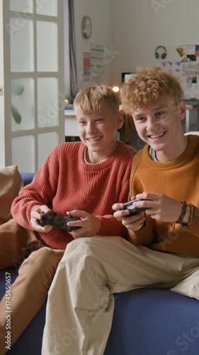 Vertical shot of two teen brothers playing videogames and nudging each other playfully fighting on sofa at home and watching TV
