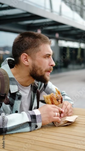 Young man with a beard and backpack enjoys eating traditional Serbian burek outdoors near a modern glass building. Vertical 4k footage