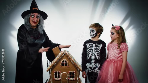 Witch with children, dressed in Halloween costumes, beside a gingerbread house, against a gray backdrop, for advertising or decoration