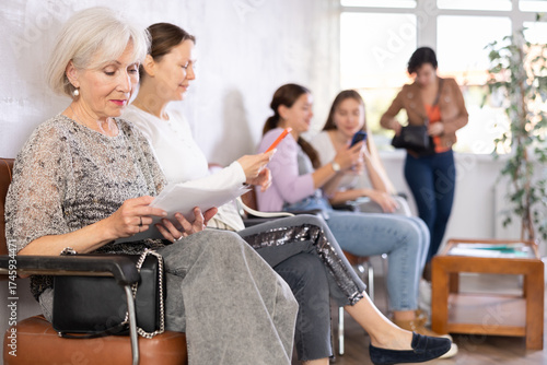 Interested senior woman sitting in lobby of clinic with papers in hand, reading medical assessment report while waiting for appointment with doctor