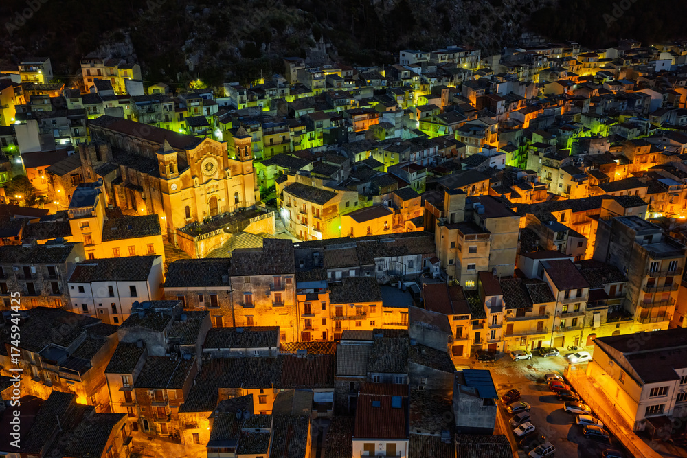 Naklejka premium Aerial drone night view of Collesano, a historic Sicilian mountain town at dusk with Basilica San Pietro church and illuminated stone streets in the Madonie Mountains, Sicily, Italy