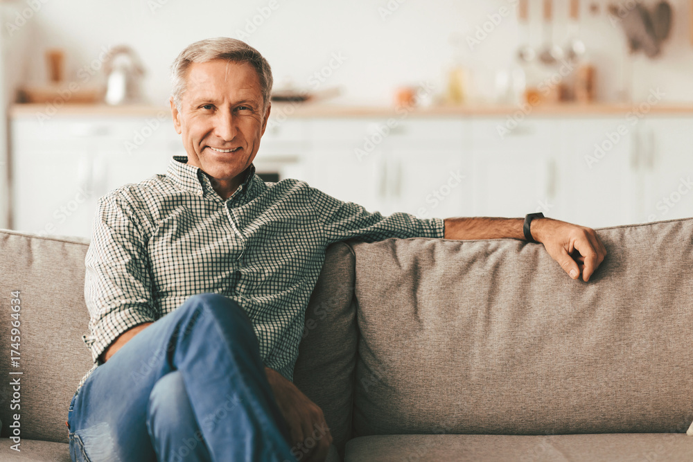 Fototapeta premium Contented Senior Man Sitting On Sofa At Home, Smiling Looking At Camera. Retired Male Relaxing On Couch In Modern Living Room Indoor On Weekend. Retirement Lifestyle, Domestic Comfort Concept