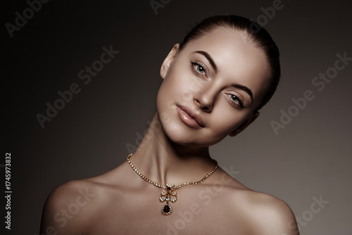 Elegant close-up portrait of a woman with nude makeup and a gold necklace with gemstone pendant. Fashion and beauty concept with soft studio lighting and neutral background.